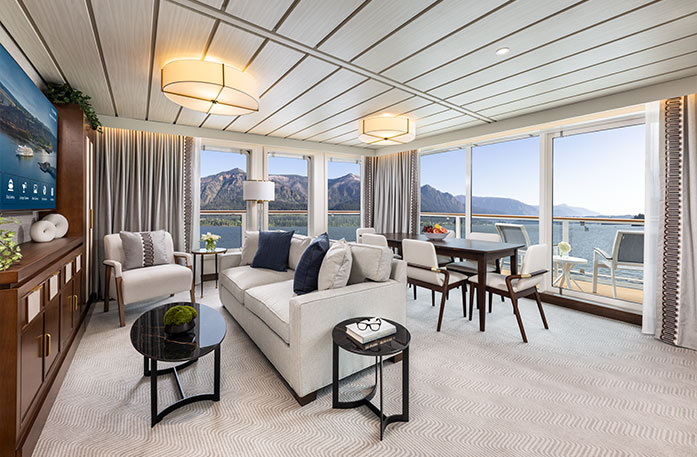 The spacious Signature Suite on American Encore, with floor-to-ceiling glass windows overlooking the private balcony and the Columbia River. There is a wooden dining table with six chairs around it, a grey sofa and an armchair, and a coffee table.