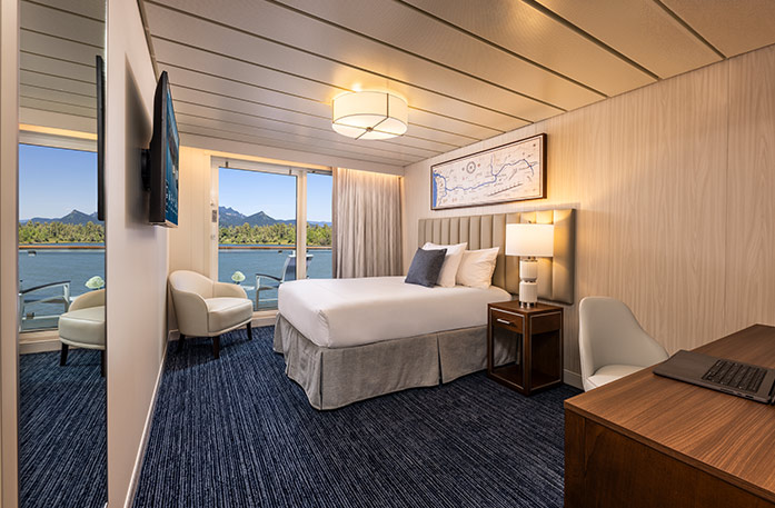 The Single Balcony stateroom on American Encore, with large floor-to-ceiling sliding glass doors, with curtains on each end, that lead to a private balcony with a table and chairs overlooking the Columbia River. There is a bed with white bedding on the right side of the room, facing a TV on the wall.