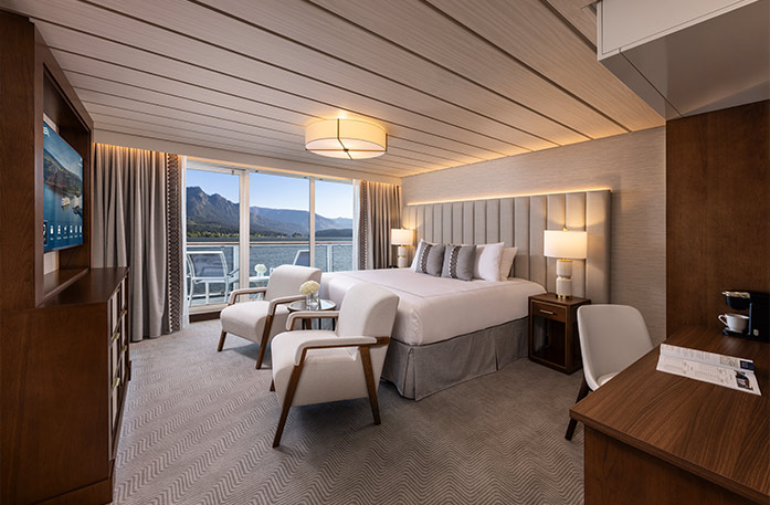 The Sky Suite on American Encore, with large floor-to-ceiling sliding glass doors leading to a private balcony overlooking the Columbia River. There is a bed against the wall on the right side with grey pillows, and two arm chairs at the foot of the bed. There is a dresser against the wall across from the bed and a TV on the wall above it.