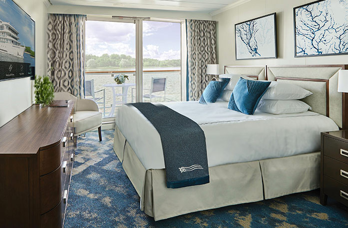 Standard Balcony stateroom on the American Harmony featuring a queen bed with blue accents, patterned curtains, a dark wood dresser, an armchair, and sliding glass doors opening to a private balcony with seating and river views.