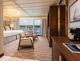 The Sky Suite on American Maverick, with large floor-to-ceiling sliding glass doors leading to a private balcony overlooking the Hudson River. There is a bed against the wall on the left side of the room, and a dresser against the wall across from the bed.