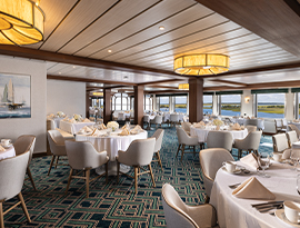 The Restaurant on American Maverick, where there are several individual dining tables surrounded by grey plush chairs in a line along a wall filled with large windows overlooking the New England coast.