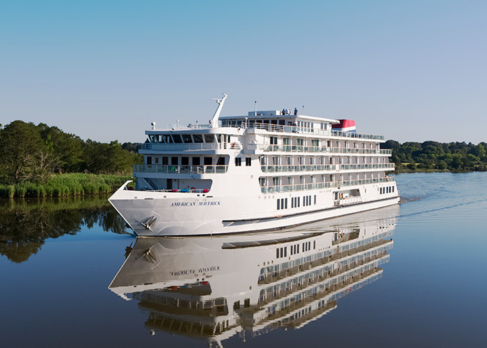 An aerial view of American Maverick, an American Cruise Lines Patriot Class small cruise ship sailing along the New England coast, with views of the shore in the distance.
