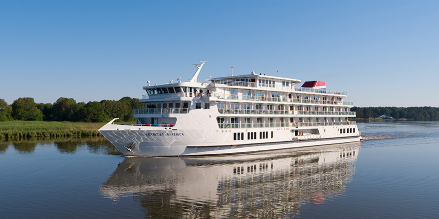 An aerial view of American Maverick, an American Cruise Lines Patriot Class small cruise ship sailing along the New England coast, with views of the shore in the distance.