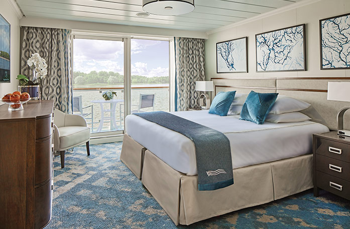 The Deluxe Balcony stateroom on American Melody, with large floor-to-ceiling glass doors with tan patterned curtains on each end that lead to a balcony with chairs overlooking the Mississippi River, a bed with white bedding and two blue pillows and a blue bed runner, and three white paintings with blue lines above the bed. There is an end table with a lamp on each side of the bed. 
