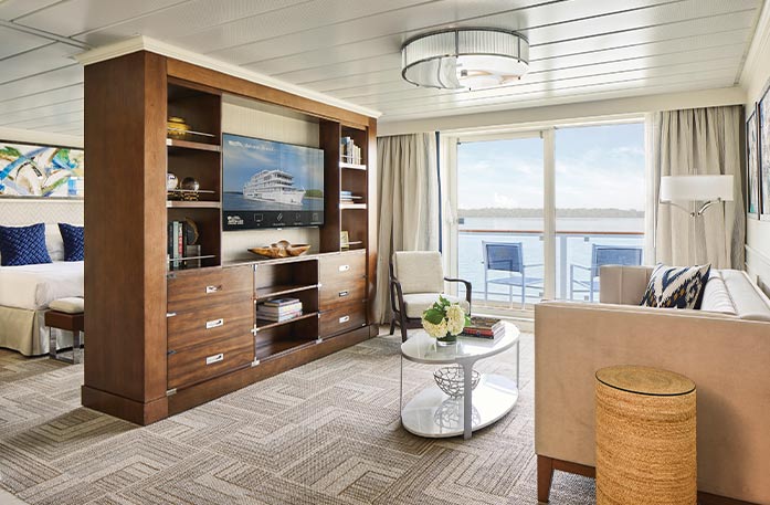 The Grand Suite on American Melody, with floor-to-ceiling glass doors that lead to a balcony with a table and chairs overlooking the Columbia River. There is a tan sofa with a black pillow beside a small table, and a large, wooden TV stand with six drawers and shelves filled with books and decor and a TV in the middle. Behind the tv stand is a bed with dark blue pillows with a painting above