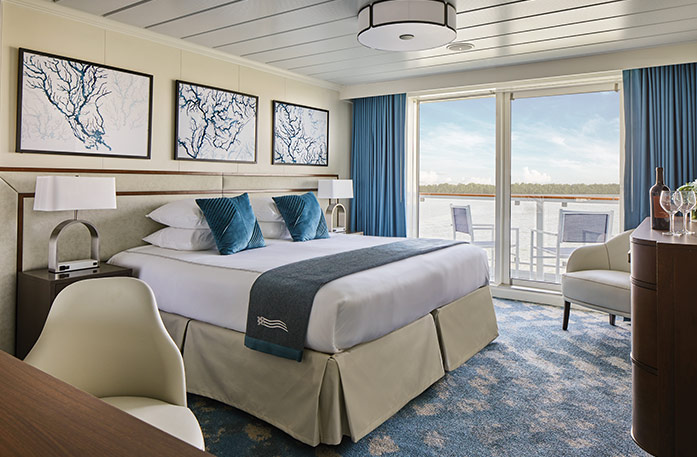 The Premium Balcony stateroom on American Melody, with large floor-to-ceiling glass doors with blue curtains on each end that lead to a balcony with chairs overlooking the Mississippi River, a bed with white bedding and two blue pillows and a blue bed runner, and three white paintings with blue lines above the bed. There is an end table with a lamp on each side of the bed. 