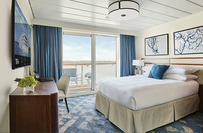The Single Balcony stateroom on American Melody, large floor-to-ceiling glass doors with blue curtains on each end that lead to a balcony with a chair and table overlooking the Mississippi River, a bed with white bedding and one blue pillow, and two white paintings with blue lines above the bed. There is a dark wood dresser and a TV on the wall in front of the bed.