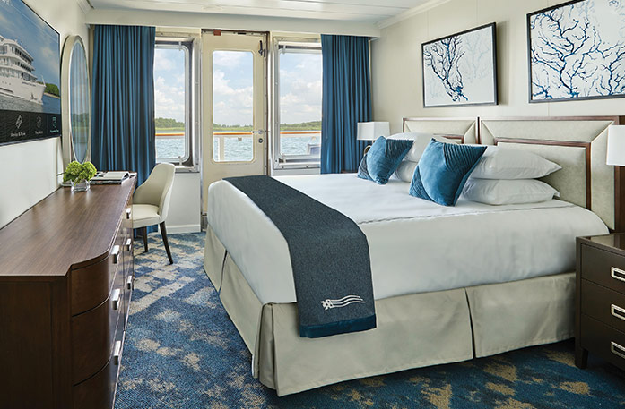 The Standard Balcony stateroom on American Melody, with a push door with blue curtains that lead to a balcony overlooking the Mississippi River, a bed with white bedding and two blue pillows and a blue bed runner, and two white paintings with blue lines above the bed. There is an end table with a lamp on each side of the bed, and a dark wood dresser and a TV on the wall in front of the bed.