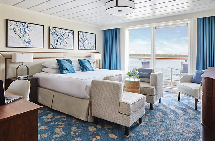 The Veranda Suite on American Melody, with large floor-to-ceiling glass doors, with blue curtains on each end, that lead to a balcony with a table and chairs overlooking the Mississippi River, a bed with white bedding and two blue pillows and two accent chairs at the foot of the bed, and three white paintings with blue lines above the bed. There is an end table with a lamp on each side of the bed.