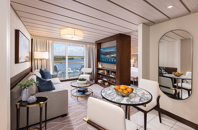 The Grand Suite on American Pioneer, with large floor-to-ceiling sliding glass doors leading to a private balcony overlooking the Florida Gulf Coast. There is a grey sofa facing a large wooden TV stand in the center of the room, with a TV and shelves with decor. 