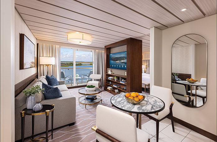 The Grand Suite on American Ranger, with large floor-to-ceiling sliding glass doors leading to a private balcony overlooking the Hudson River. There is a tan sofa facing a large wooden TV stand in the center of the room, with a TV and shelves with decor. 