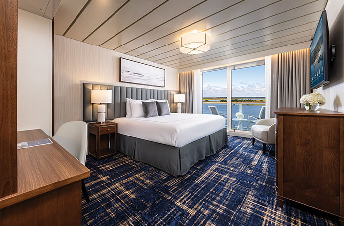 The Premium Balcony stateroom on American Ranger, with floor-to-ceiling sliding glass doors that lead to a private balcony overlooking the Hudson River. There is a bed against the wall on the left side of the room with a blue bed runner and accent pillows, an end table with a lamp on each side, and three paintings above the bed.