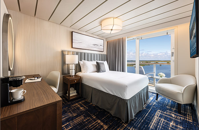The Single Balcony stateroom on American Ranger, with floor-to-ceiling sliding glass doors that lead to a private balcony overlooking the Hudson River. There is a bed against the wall on the left side of the room with a blue bed runner and a grey accent pillow, an end table with a lamp on one side, and a dresser on the wall across the room with a TV above it.