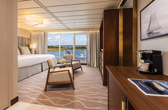 The Sky Suite on American Ranger, with large floor-to-ceiling sliding glass doors leading to a private balcony overlooking the Hudson River. There is a bed against the wall on the right side of the room with navy blue pillows, tan bedding, a grey bed runner, and a navy blue and gold bench at the foot of the bed. There is a dresser against the wall across from the bed and a TV on the wall above it.