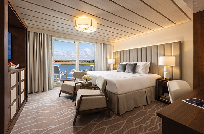 The Veranda Suite on American Ranger, with floor-to-ceiling sliding glass doors that lead to a private balcony overlooking the Hudson River. There is a bed against the wall on the left side of the room with a blue bed runner and accent pillows, an end table with a lamp on each side, and three paintings above the bed.