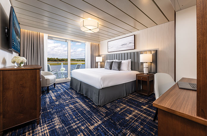 The Vista Balcony stateroom on American Ranger, with floor-to-ceiling sliding glass doors that lead to a private balcony overlooking the Hudson River. There is a bed against the wall on the left side of the room with a blue bed runner and accent pillows, an end table with a lamp on each side, and three paintings above the bed.