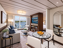 The Veranda Suite on American Ranger, with floor-to-ceiling sliding glass doors that lead to a private balcony overlooking the Hudson River. There is a bed against the wall on the left side of the room with a blue bed runner and accent pillows, an end table with a lamp on each side, and three paintings above the bed.