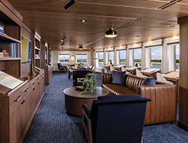 The Restaurant on American Ranger, with several tables and chairs perfectly placed on the white and green patterned carpet. There is a mural on the back wall, as well as large windows overlooking the Hudson River.