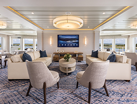 The Chesapeake Lounge, a common area on American Ranger, with two tan sofas, several lounge chairs and small round tables, and a small bar in the back corner. The right wall is filled with large windows overlooking the sunset on the Hudson River.