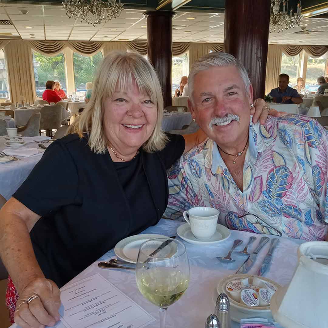 Smiling couple seated at a table in a cruise ship dining room with large windows showing scenic river view in the background.