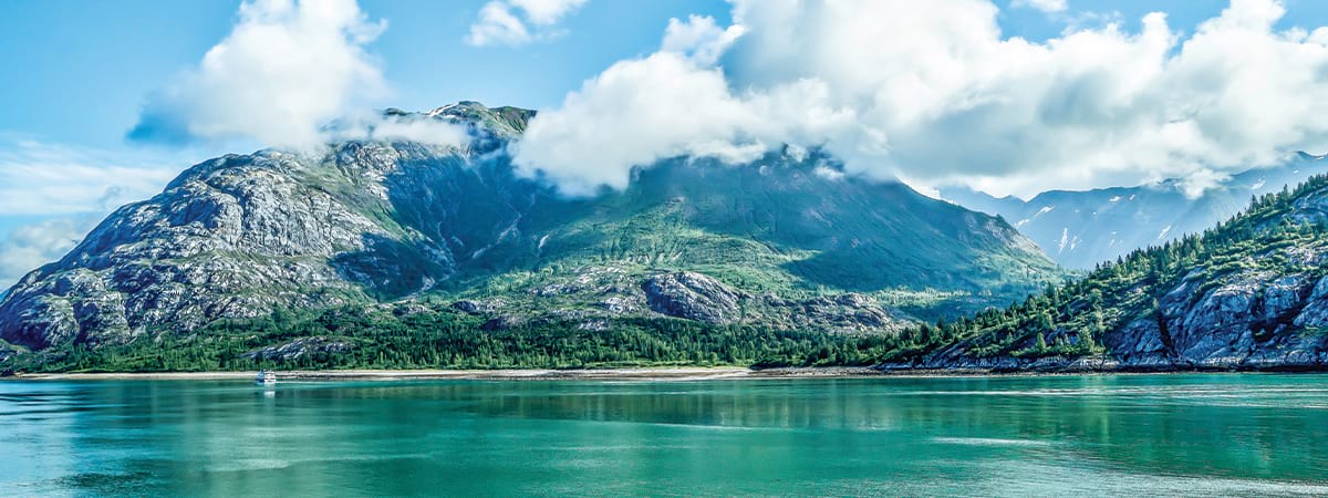Small American Cruise Lines ship on calm river, with green covered mountains in the background, under a blue sky with clouds.