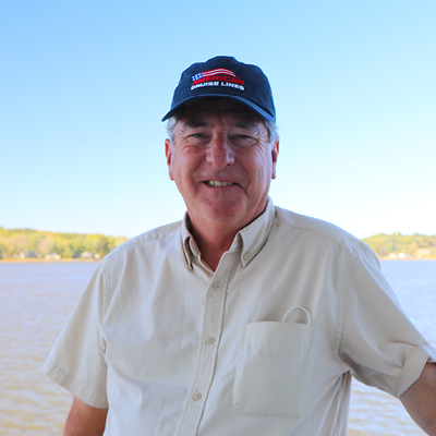 Jim Zim, an American Cruise Lines guest and social media influencer, smiling for a photo with the Tennessee River in the background. He is wearing a navy blue cap with the American Cruise Lines logo on the front, and a yellow button-down t-shirt.