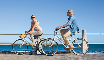 A man and a woman riding bikes alongside a body of water while smiling at each other.