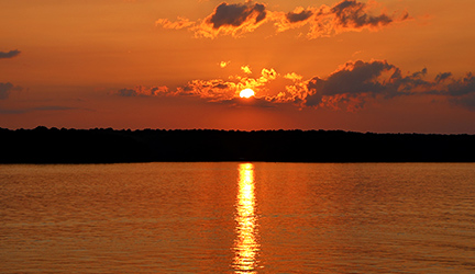 A breathtaking sunset creates an orange glow in the sky, reflecting over the waters of the Tennessee River. There is a dark silhouette of mountains in the distance.