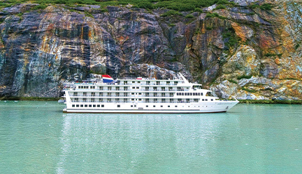 American Constellation, an American Cruise Lines small ship, sailing along the teal waters of Alaska the beautiful Alaskan cliffs in the background.