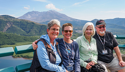 A group of friends gathered together, posing for a picture in front of tall mountains in the distance while aboard a cruise.