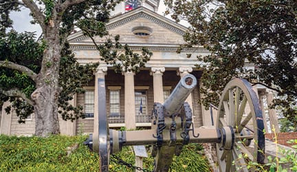 A cannon with two large wheels is pictured in front of the historic Old Warren County Courthouse, a light brick building with pillars out front in Vicksburg.