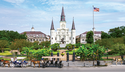 The St. Louis Cathedral with three spires stands in Jackson Square in New Orleans, while a statue of a rider on a horse is centrally placed in a manicured park with trees and bright greenery, and horses drop off carriages of visitors in the front. 