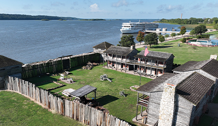 In the distance, an American Cruise Lines modern riverboat is docked at a port long the Mississippi River near a small town and historic battlefield that is surrounded by a jagged wooden fence.