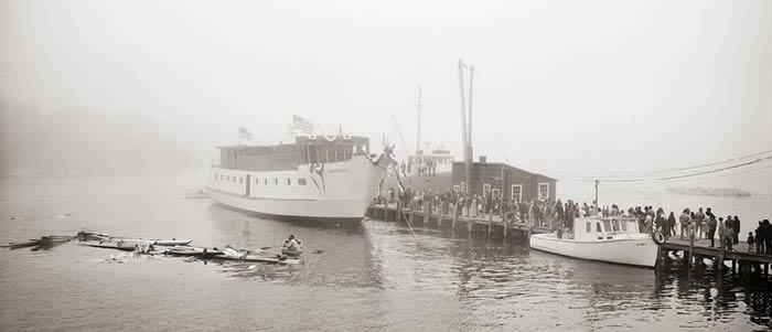 Small ship docked with passengers boarding and rowers in water with a foggy background.