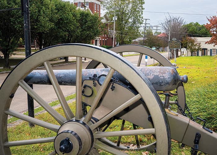 A close up view of a cannon with two large wheels on each side, with a view of a tall brick building in the distance in Vicksburg.