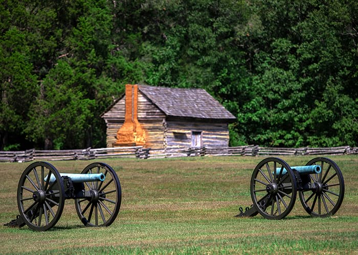 Two cannons, each with two large wheels, in the middle of the historic Shiloh National Military Park. In the distance, there is a small log cabin with a large chimney along the side and a long wooden fence in front.
