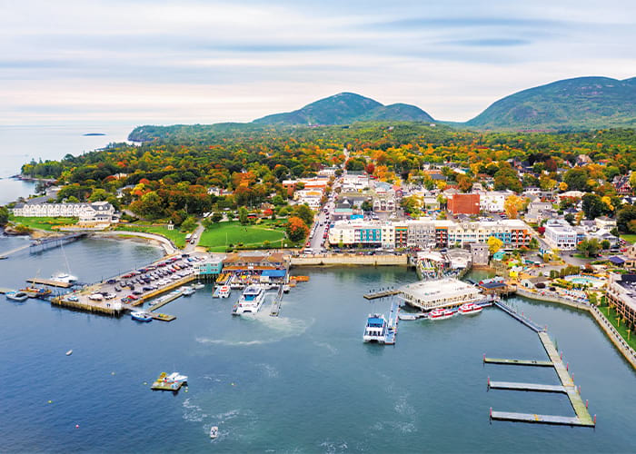 An aerial view of Bar Harbor, a town along the water in Maine with several boats docked in the marina, buildings by the shore, and countless fall foliage trees. There are small mountains in the distance along the horizon.