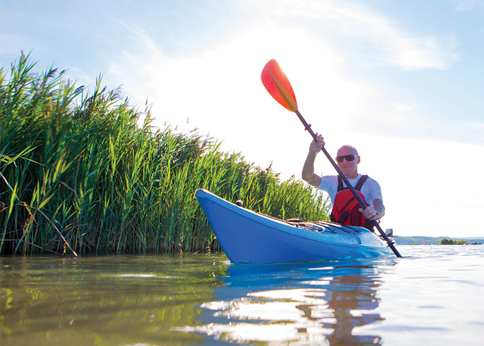 A man in a bright blue kayak with a black and orange paddle, sailing along Pool 6 on the Mississippi River in Winona, beside tall grasslands coming out from the water.