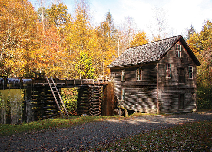 A historic, two-story log cabin in Cades Cove, a valley within Great Smoky Mountains National Park. To the left of the cabin, there are logs carefully crafted as support beams beneath a water mill that leads into a small waterfall streaming from it. The tall trees of the Great Smoky Mountains are in the distance, with their orange and red fall foliage leaves scattered along the ground.