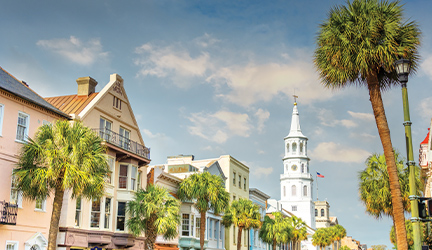 An endless line of colorful buildings and tall palm trees along a street in Downtown Charleston, including the tall, white spire on top of St. Michael's church.