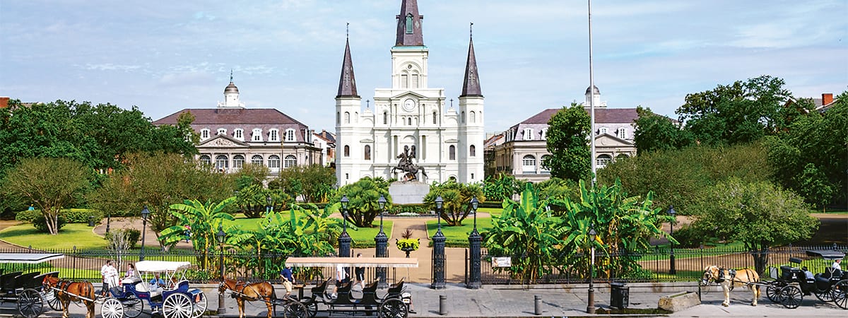 The St. Louis Cathedral with three spires stands in Jackson Square in New Orleans, while a statue of a rider on a horse is centrally placed in a manicured park with trees and bright greenery.