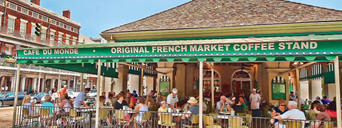 The French Market in New Orleans, a large outdoor dining area attached to a building, where there are dozens of people sitting at the tables. There is a green roof covering the dining area, with Cafe Du Monde and Original French Market Coffee Stand written along the top.