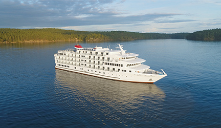 A small white cruise ship sailing on beautiful blue water surrounded by bright green forests..
