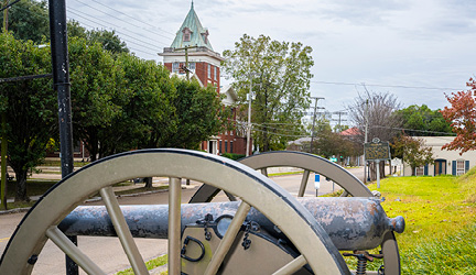 A close up view of a cannon with two large wheels on each side, with a view of a tall brick building in the distance in Vicksburg.