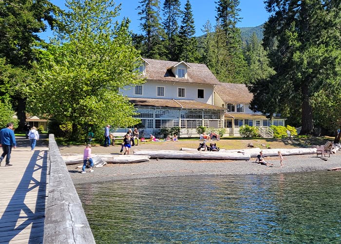 A bridge guides people over a body of water leading to a house surrounded by bright green trees and several visitors enjoying the view.