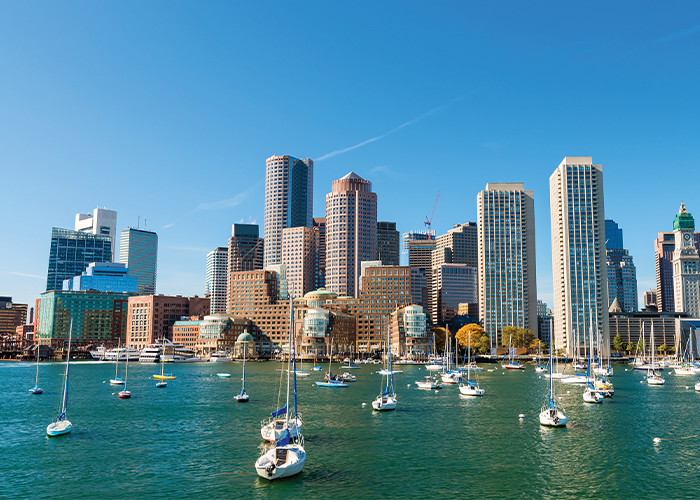 A scenic landscape of boats along the Boston Harbor, with numerous buildings and skyscrapers along the horizon line in Boston.