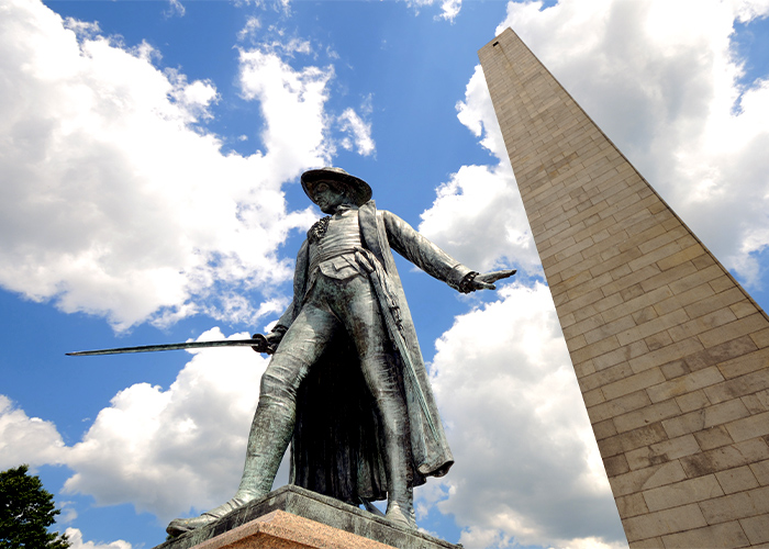 A bottom-up view of the Colonel William Prescott Statue, a bronze statue of a William Prescott wearing a wide-brimmed hat and holding a sword beside the tall, brick Bunker Hill monument along the Freedom Trail in Boston.
