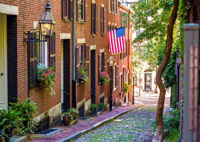 A narrow cobblestone street on a hill lined with a large, brick buildings on the left side and a brick sidewalk in Beacon Hill. The buildings have several window and ground planters with bright colored flowers, black street lamps, and an American flag flying from a second story window.