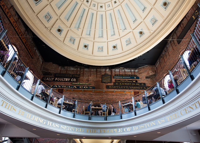 A bottom-up view of the inside of Faneuil Hall Marketplace, where there is an oval-shaped balcony with railings and tables filled with people sitting around them, and a tan dome roof with silver detailing.